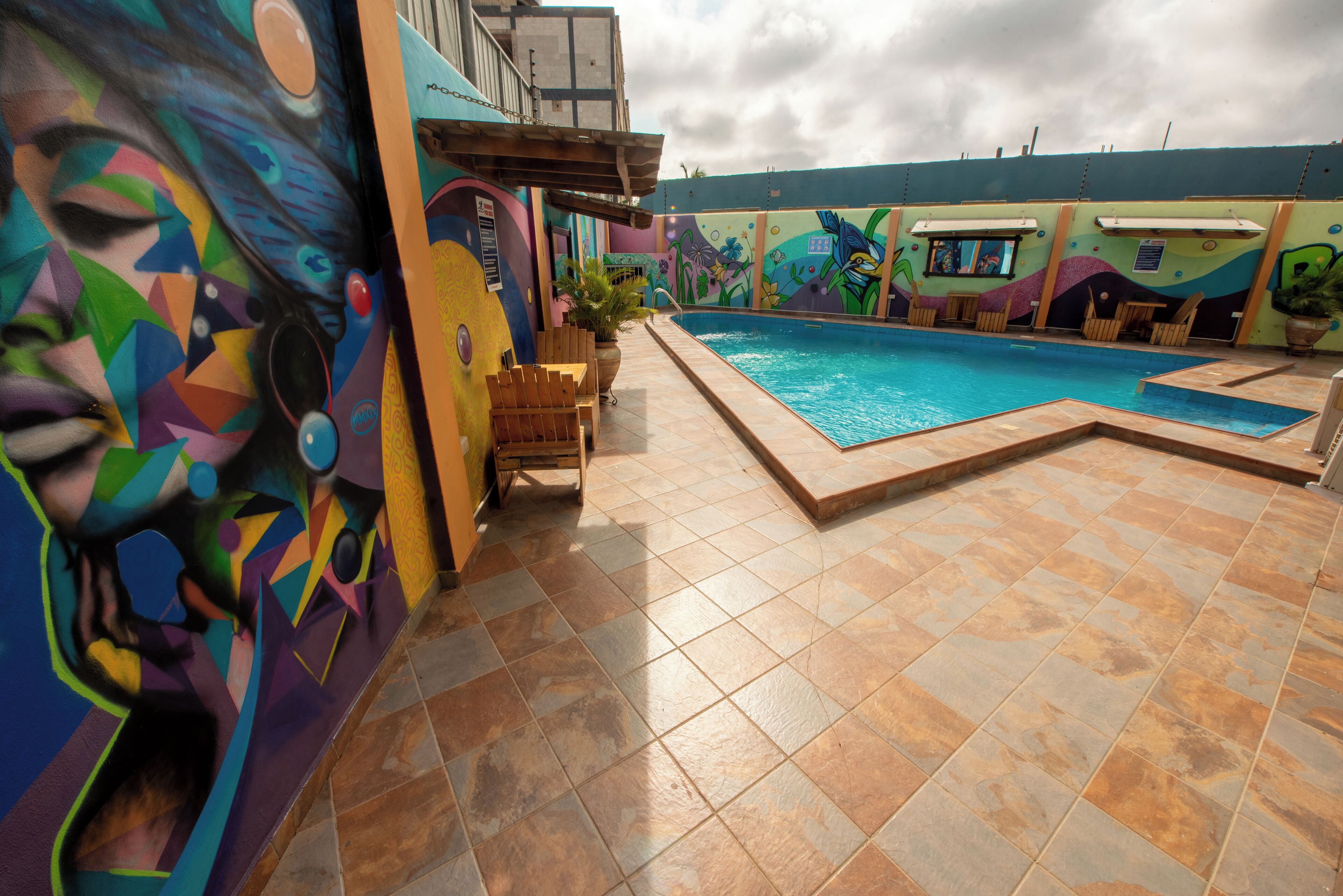 A swimming pool area with colorful murals, tiled flooring, and wooden chairs.