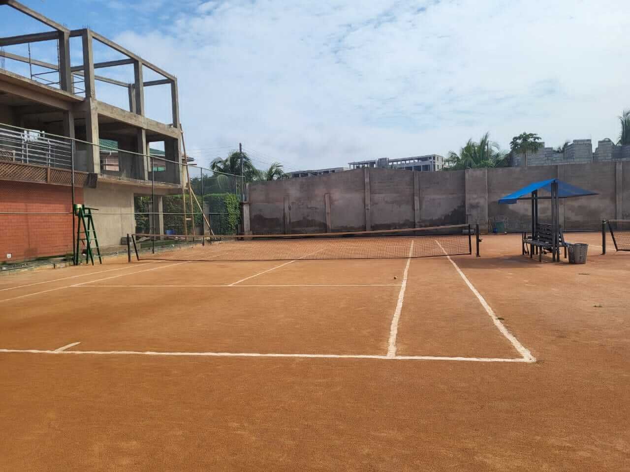 Una cancha de tenis interior con superficie de arcilla, rodeada por un muro alto y un edificio con balcón.