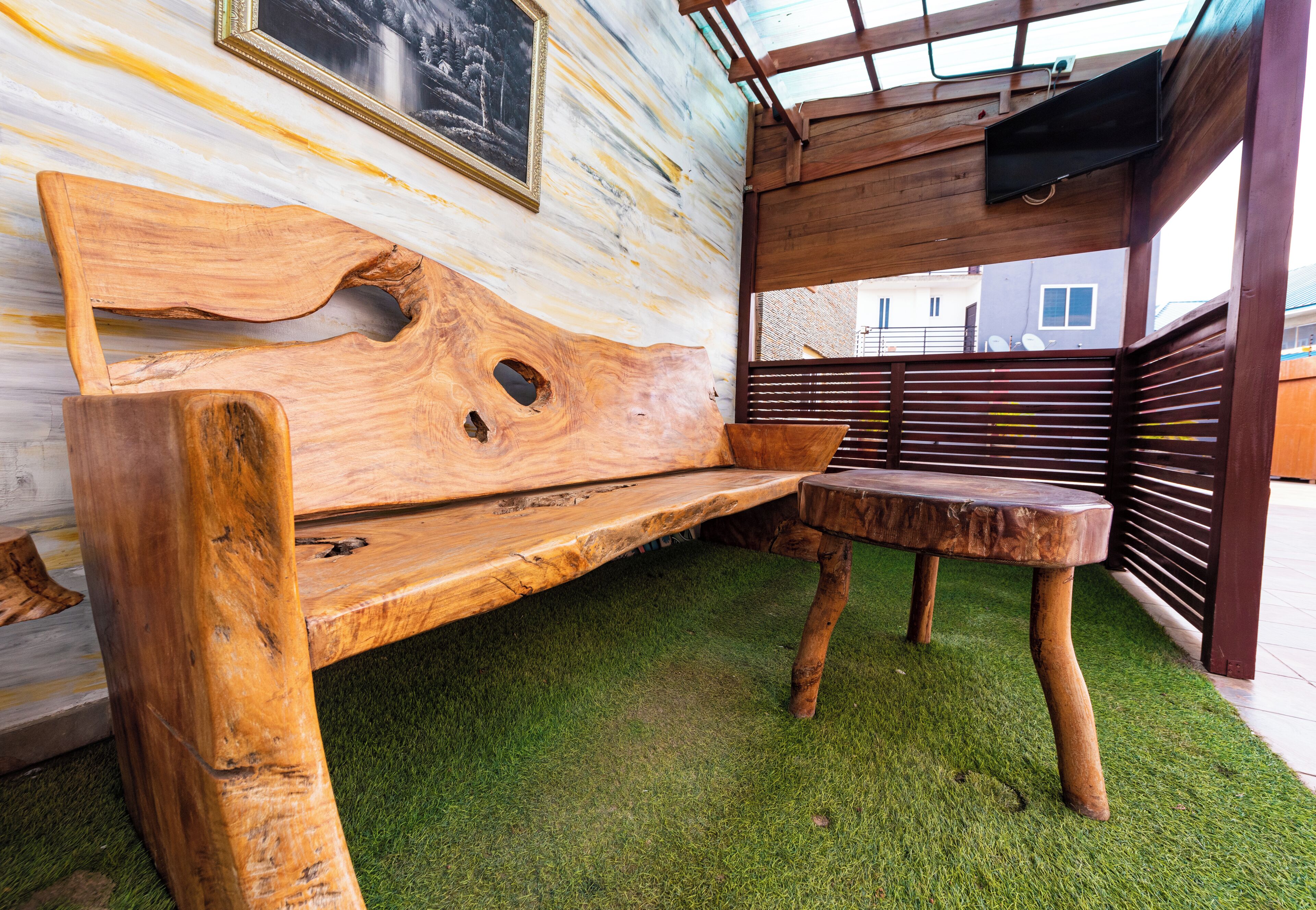 A wooden bench and table set on artificial grass in a covered outdoor area with a wooden ceiling and a wall-mounted picture frame.
