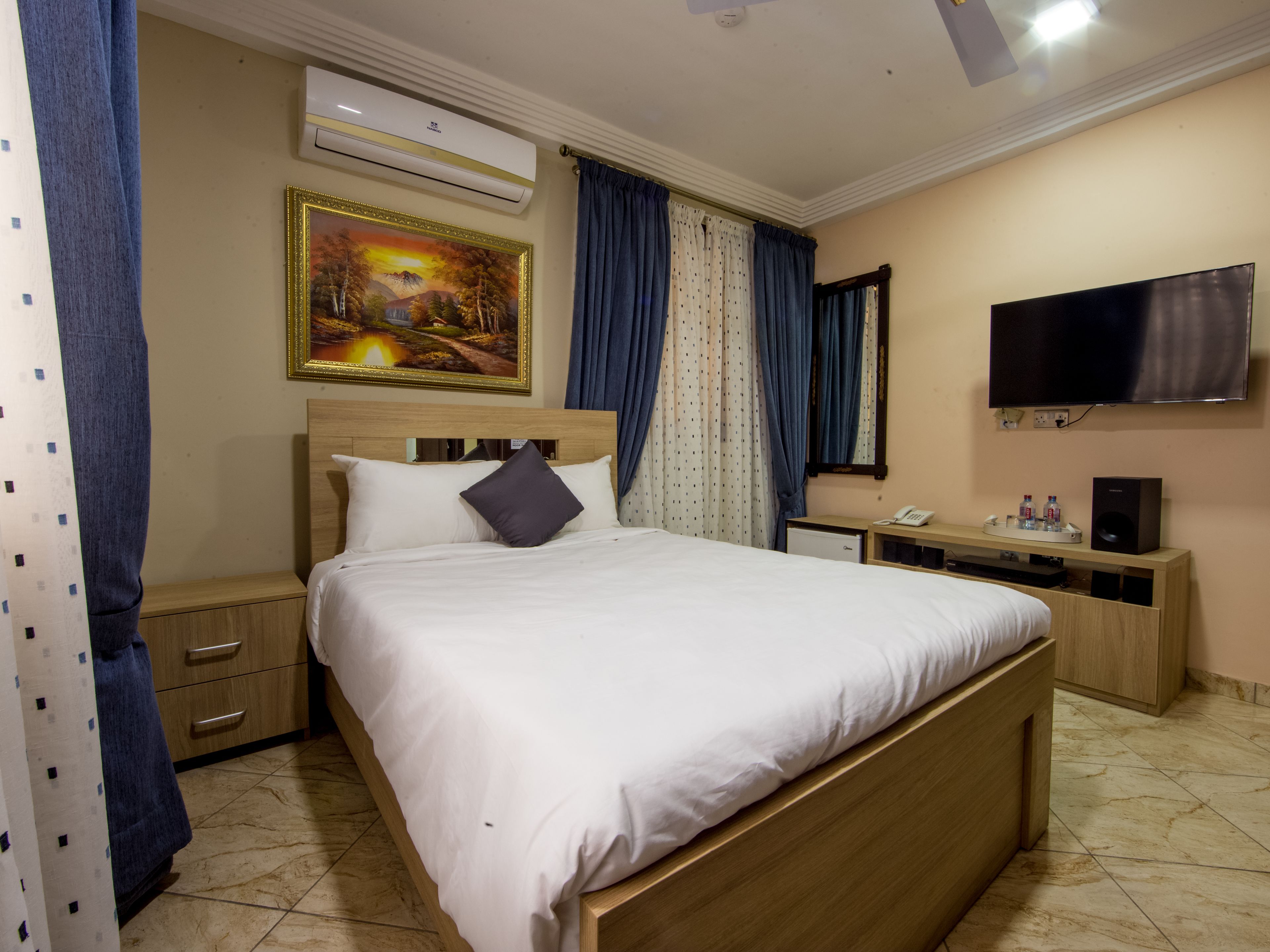 Habitación de hotel con cama, escritorio con teléfono y altavoz, televisión, aire acondicionado y un cuadro en la pared.