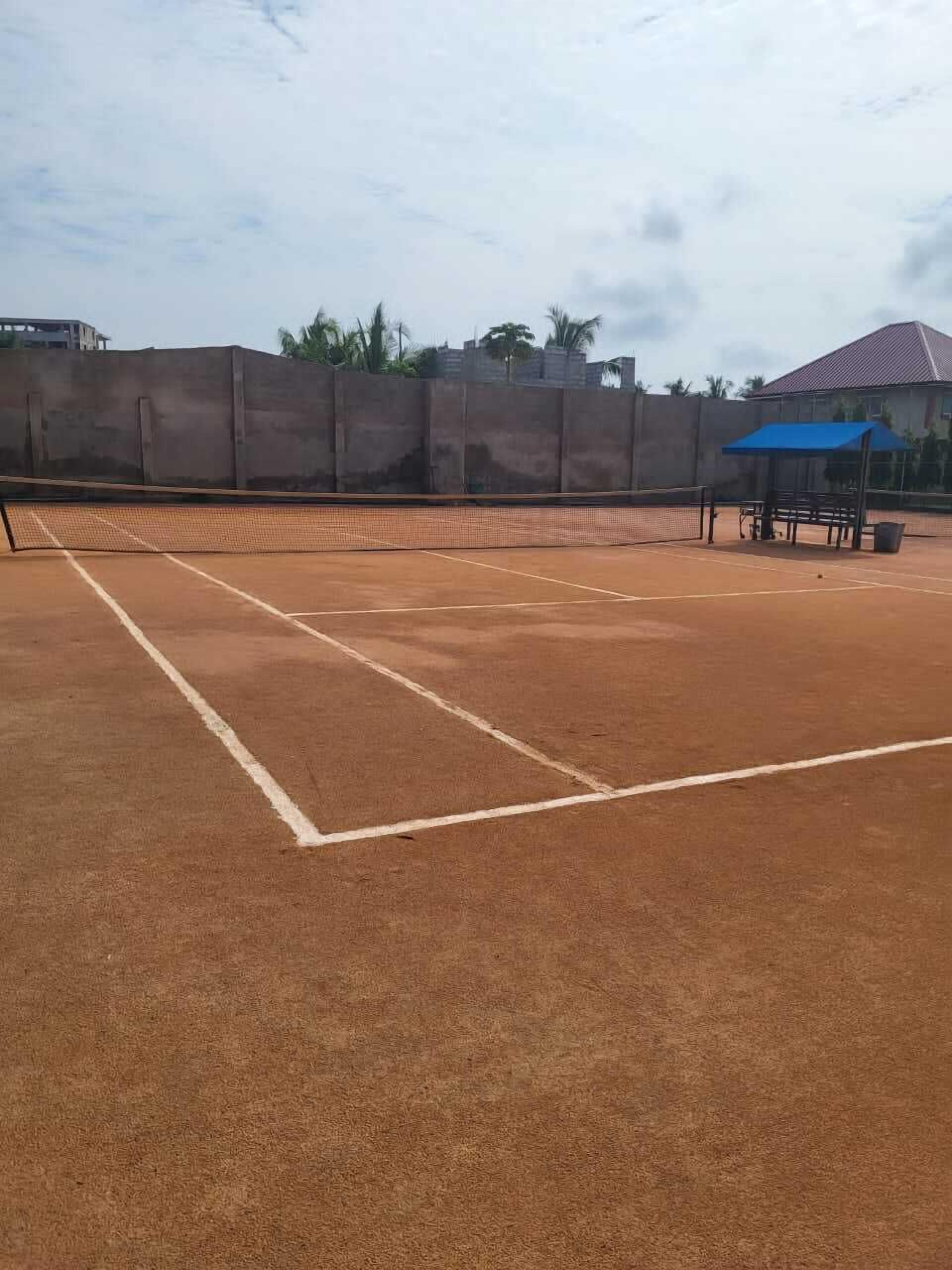 An outdoor tennis court with a net, surrounded by a high wall and a blue-roofed pavilion.