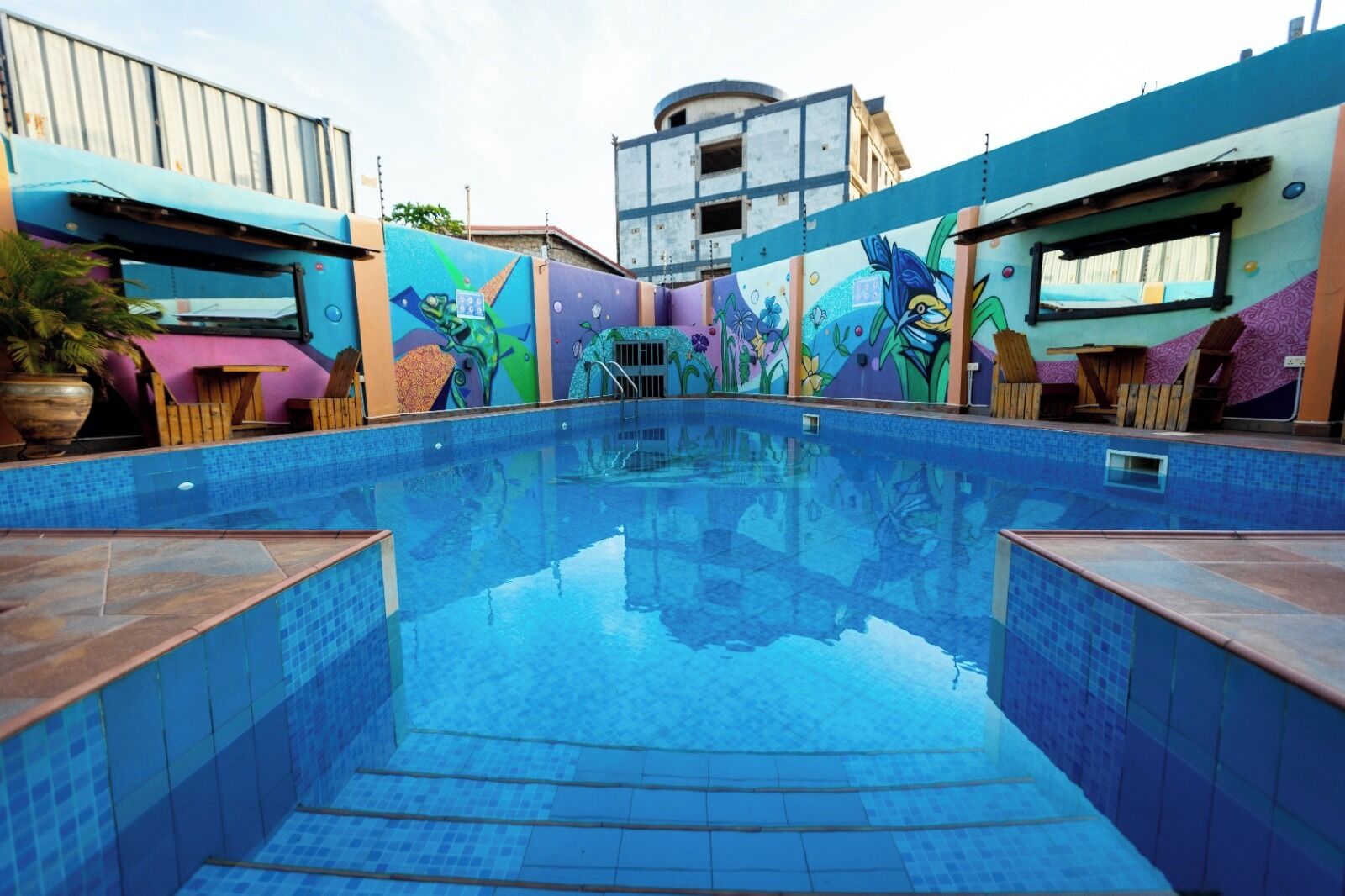 An outdoor swimming pool with a colorful mural on the walls, surrounded by wooden furniture and potted plants.