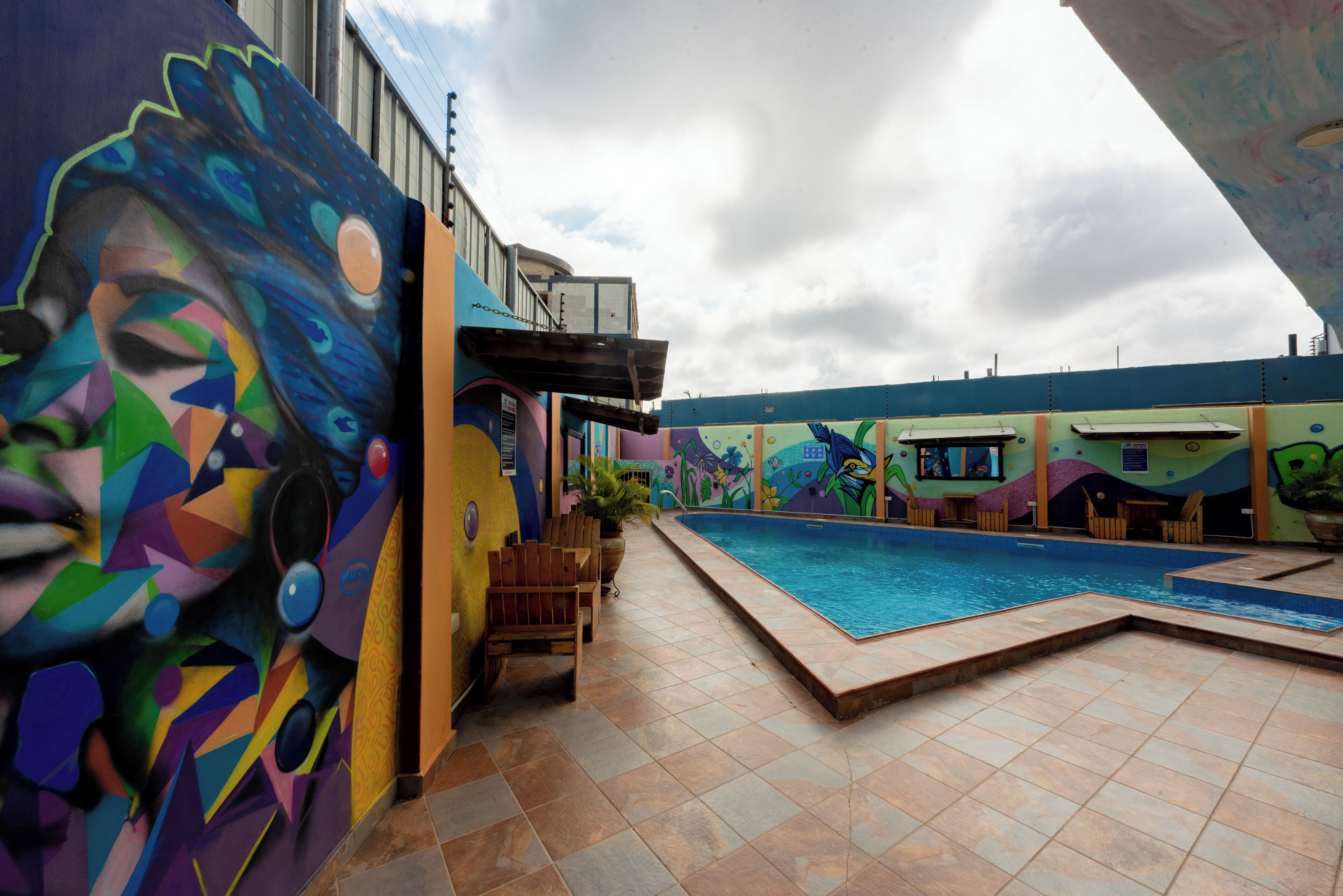 A swimming pool area with colorful murals on the walls, wooden chairs, and a tiled floor.