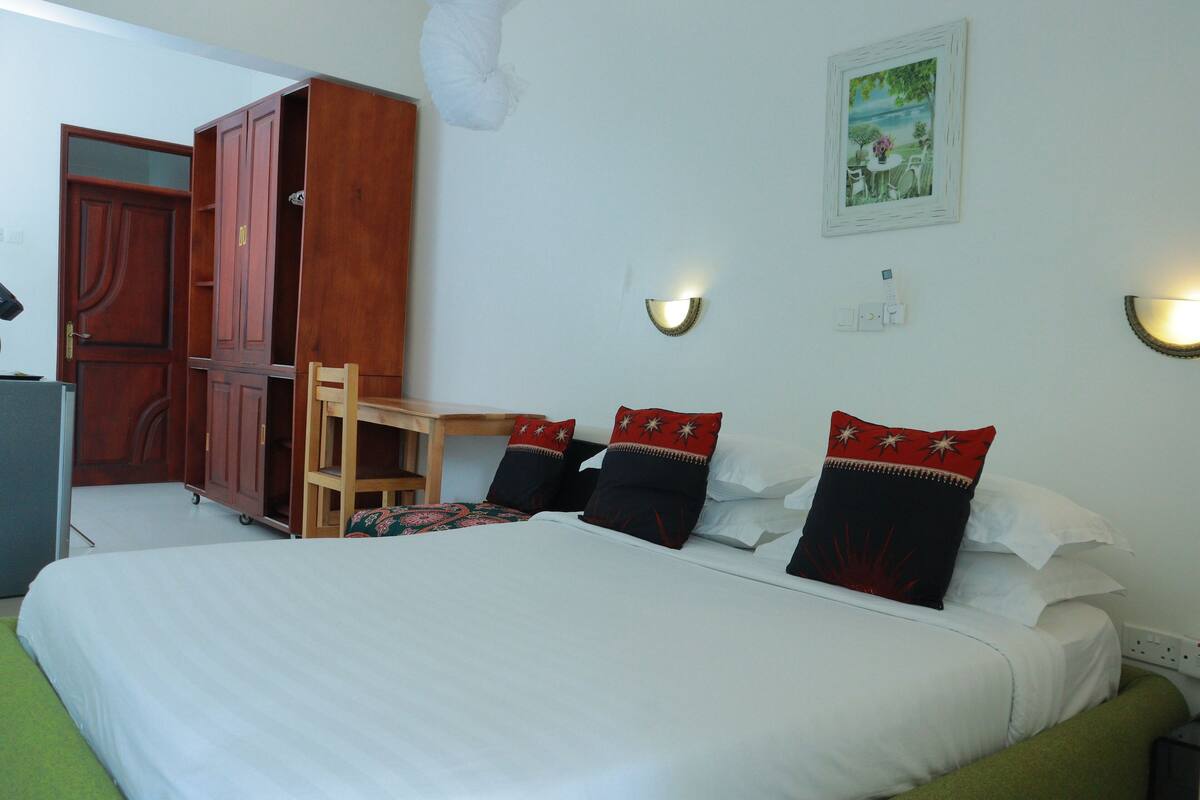 A hotel room with a large bed, a wooden wardrobe, a desk, and two wall-mounted lights.