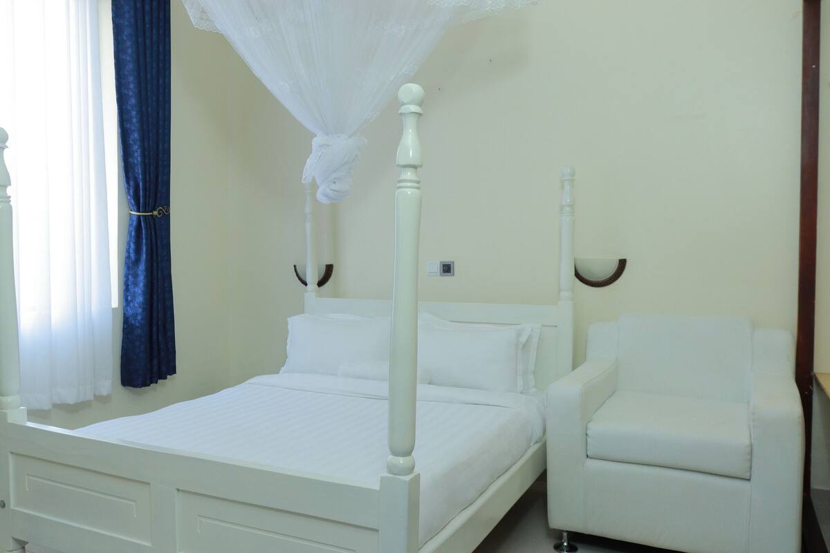 A white four-poster bed with a canopy, a white armchair, and a bedside table in a room with cream walls.