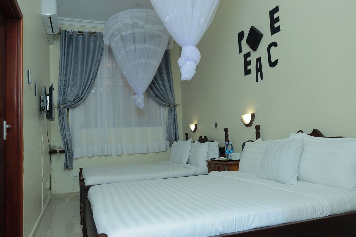 A hotel room with two beds, a wall-mounted air conditioner, a bedside table with a lamp, and a wall with a decorative sign.