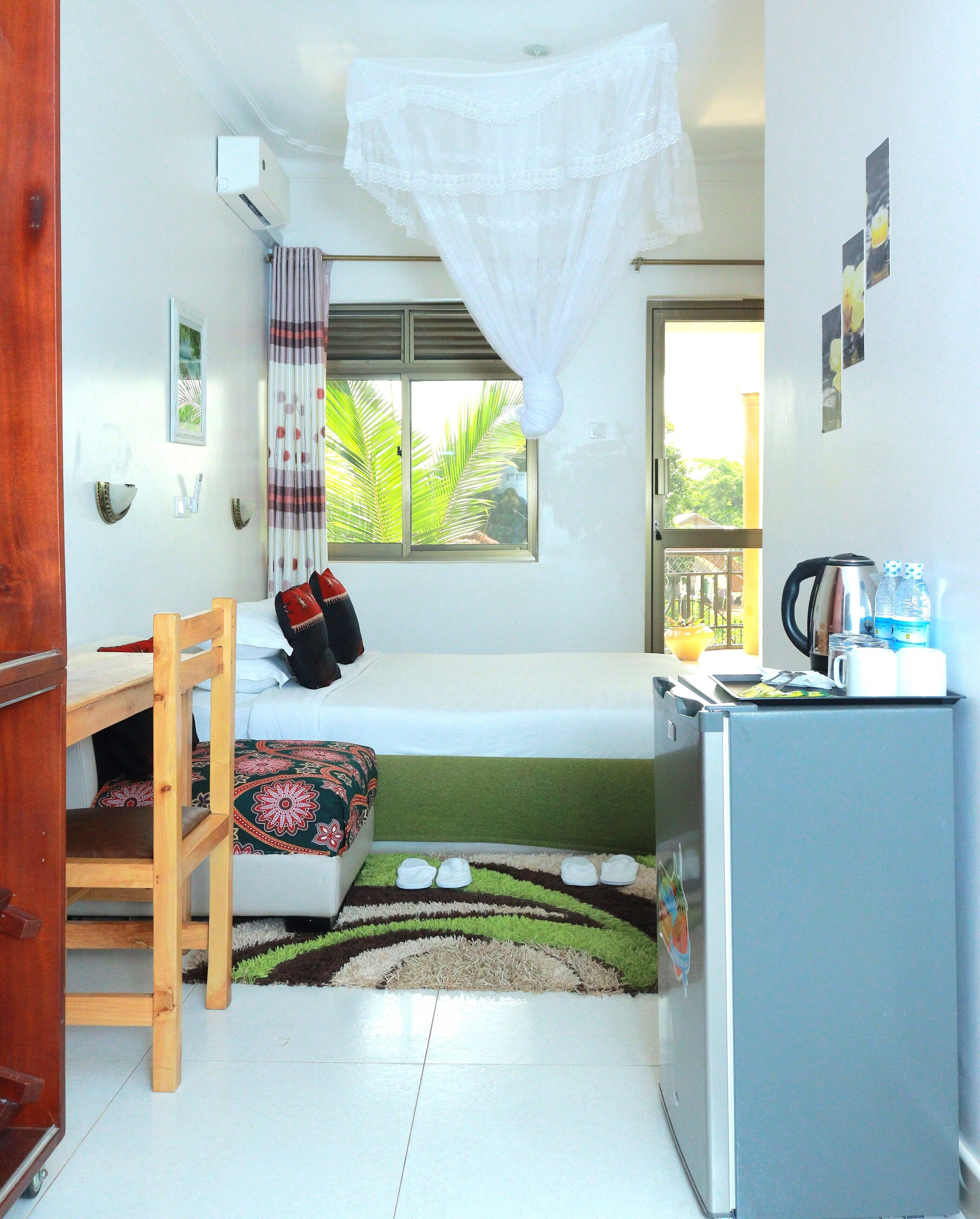 A compact room with a bed, a small table, a refrigerator, and a window with a view of greenery.