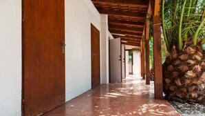 A corridor with wooden doors and a tiled floor, leading to a covered area with a wooden ceiling and a stack of coconuts. - Villaggio Riccio (Mattinata)