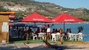 A beachside restaurant with outdoor seating and red umbrellas, overlooking a coastal area with hills in the background. - Villaggio Riccio (Mattinata)