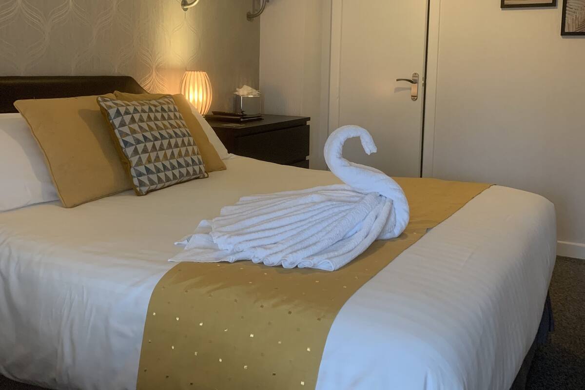 Quarto de hotel com cama bem arrumada, mesa de cabeceira com abajur, toalheiro fixado na parede e uma porta.