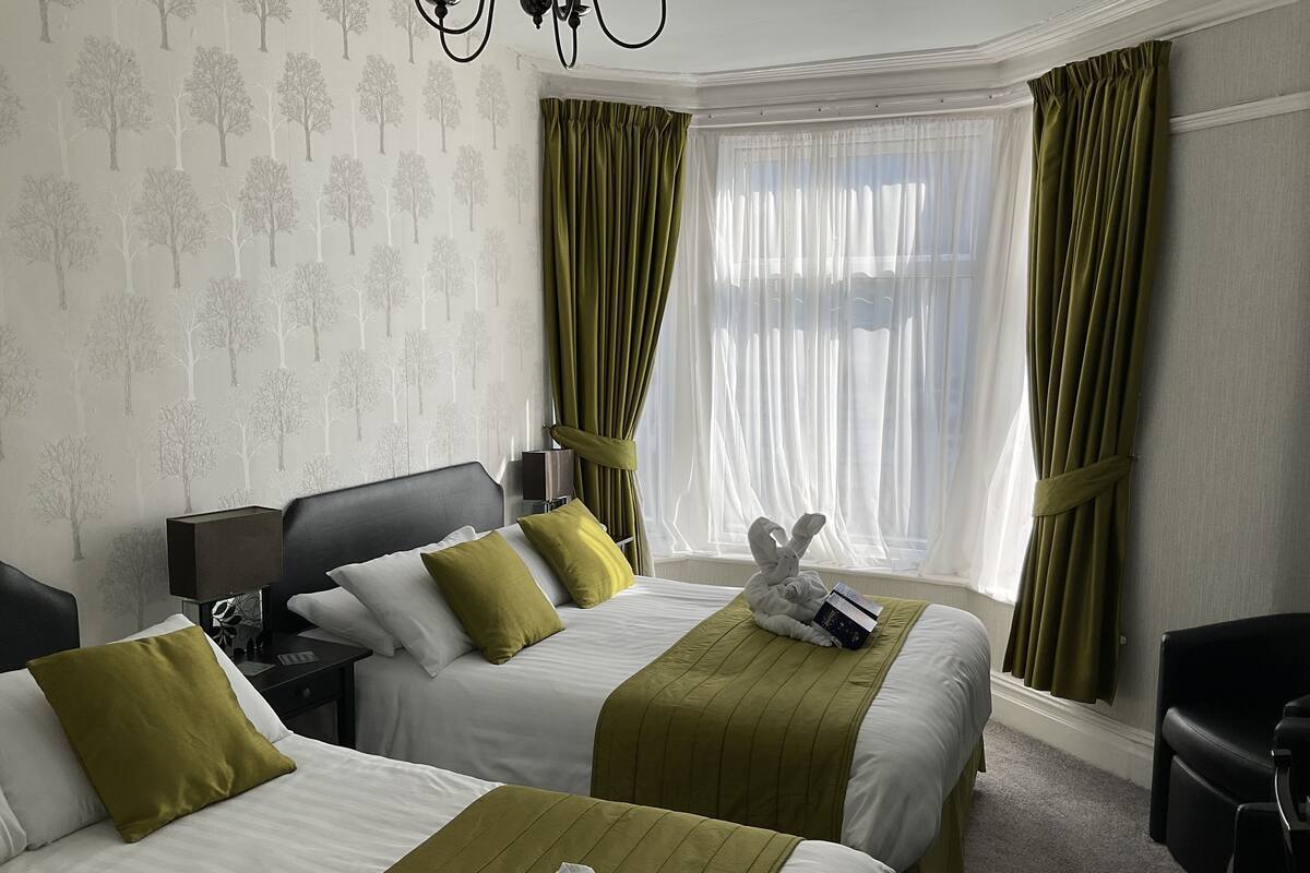 Quarto de hotel com duas camas, uma mesinha de cabeceira, uma luminária, uma cadeira e uma janela com cortinas leves.