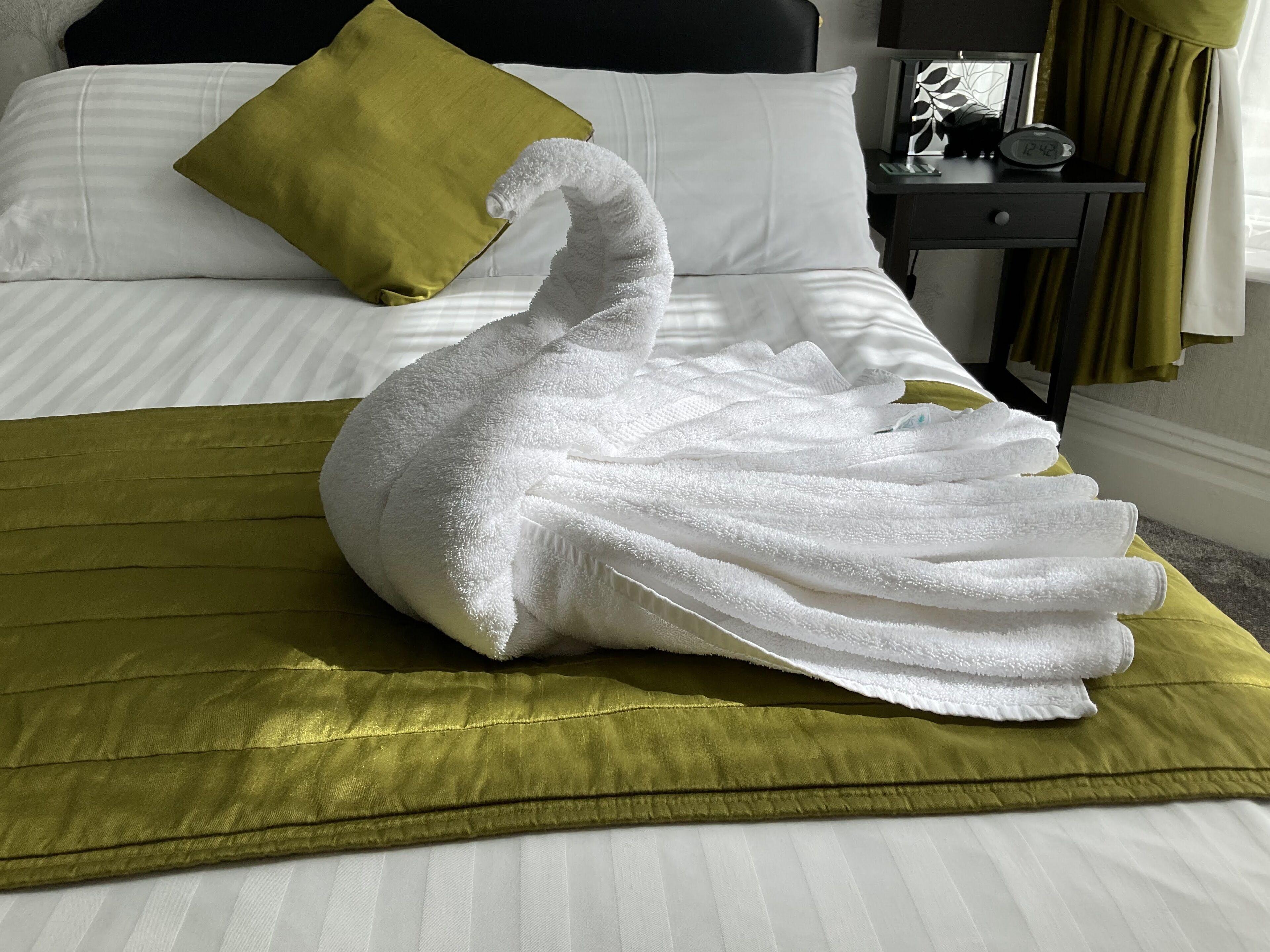 Una cama con un albornoz blanco con forma de cisne, una almohada verde y una mesita de noche con un reloj.