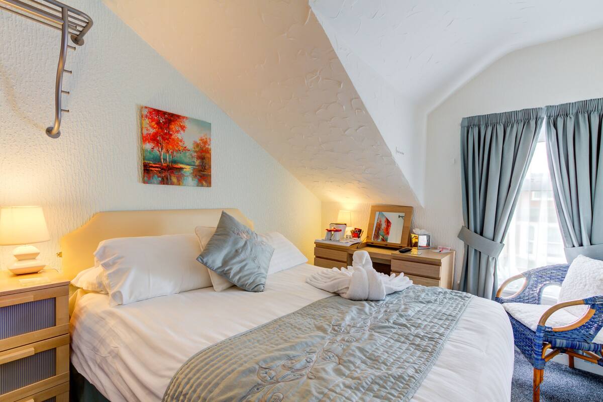 Quarto de hotel com cama arrumada, mesa de cabeceira com abajur, quadro na parede e janela com cortinas.