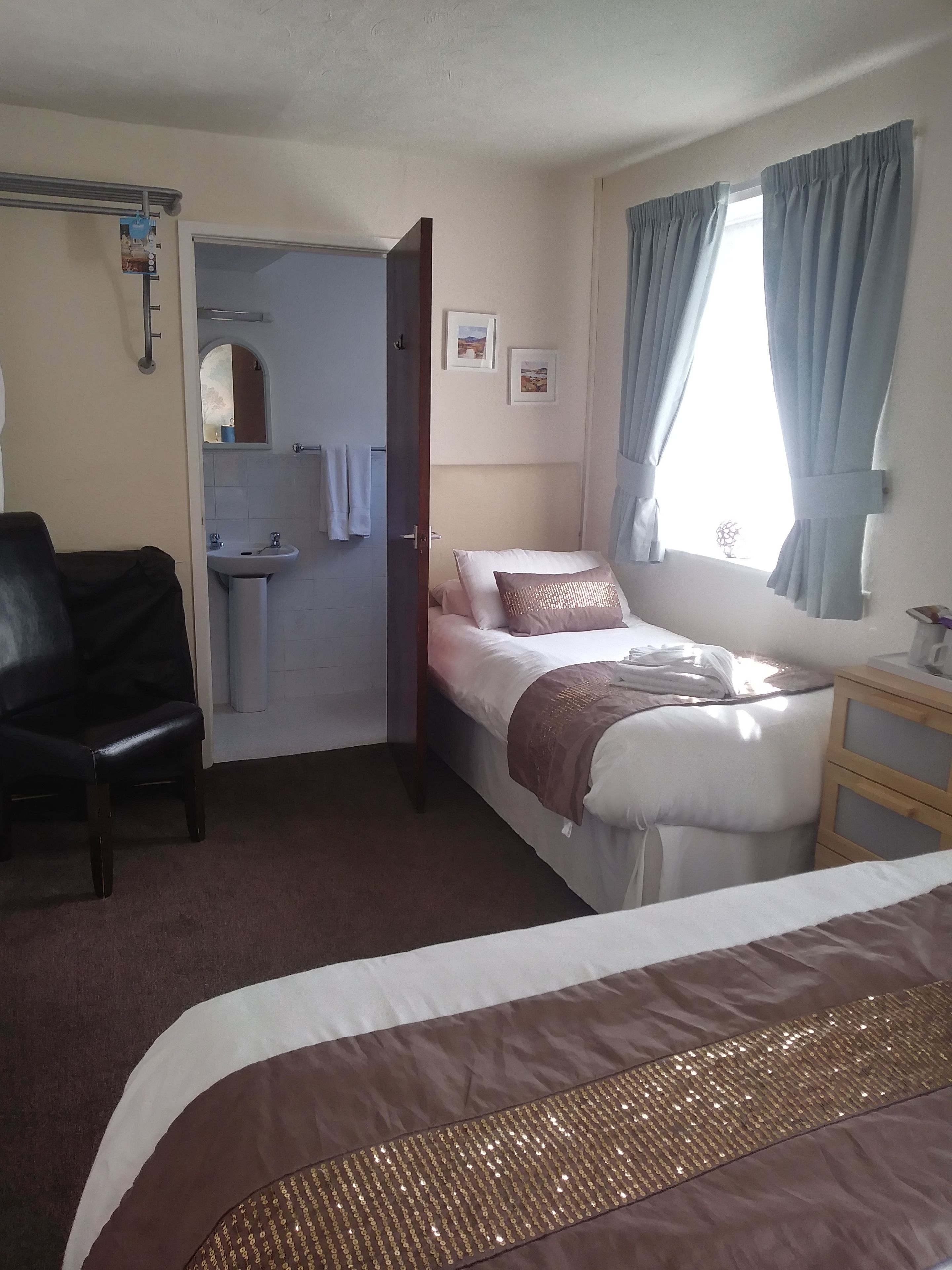 A hotel room with a bed, a chair, a small table, a window with curtains, a sink, and a mirror.