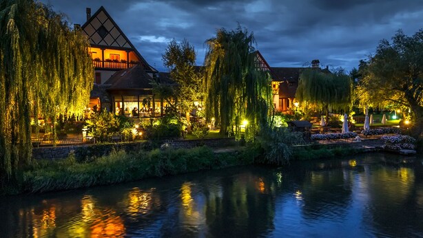 A traditional European-style building with a prominent gabled roof and a well-lit garden area adjacent to a calm water body.
