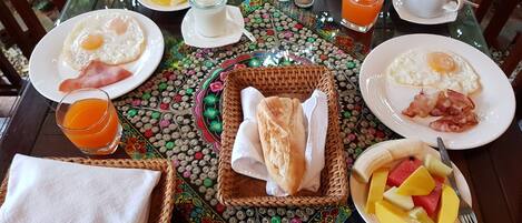 A breakfast table with plates of eggs, bacon, and fruit, a teapot, glasses of orange juice, and a basket of bread.