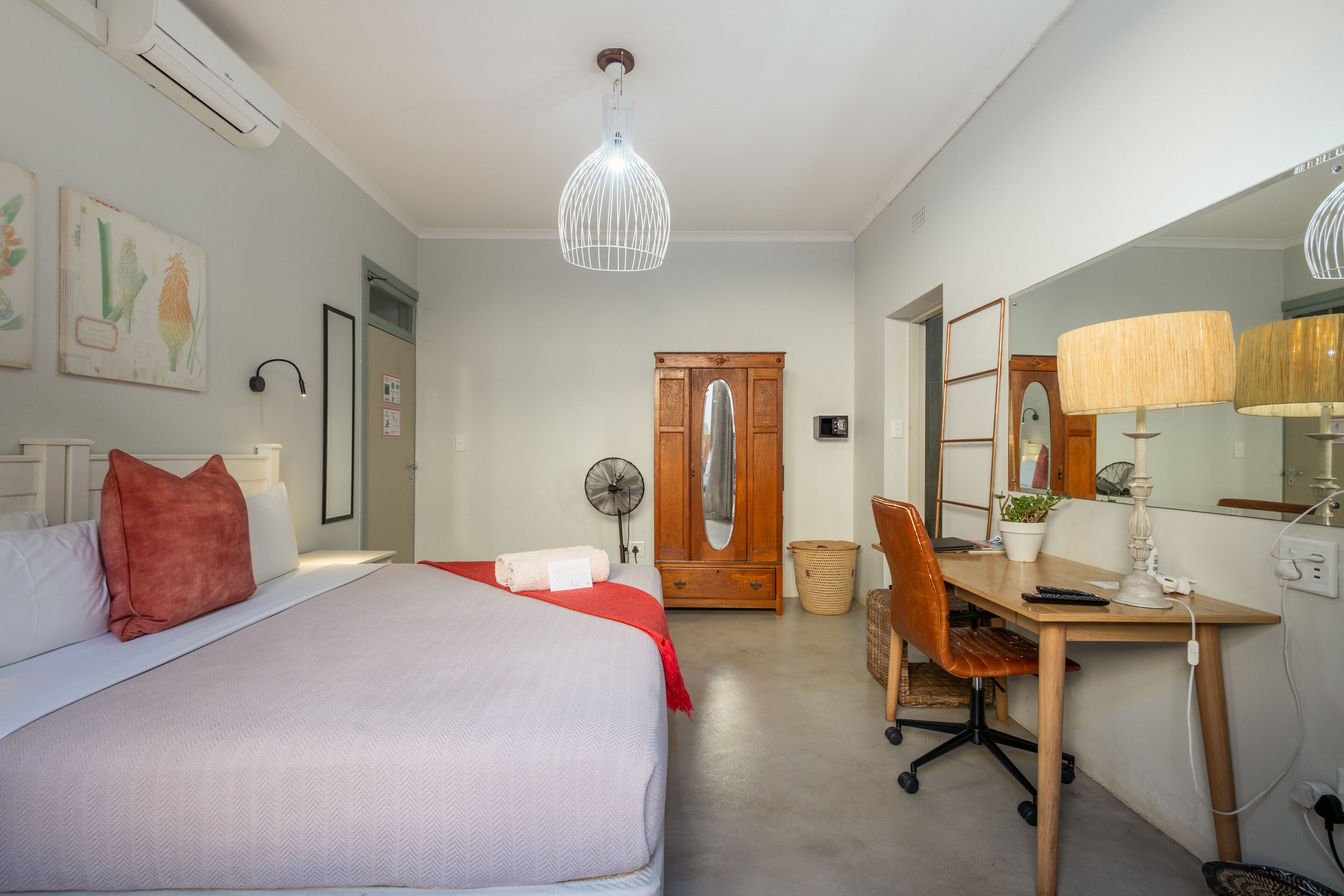 Un dormitorio con una cama, un escritorio y una silla. Hay un espejo, una lámpara y un ventilador.