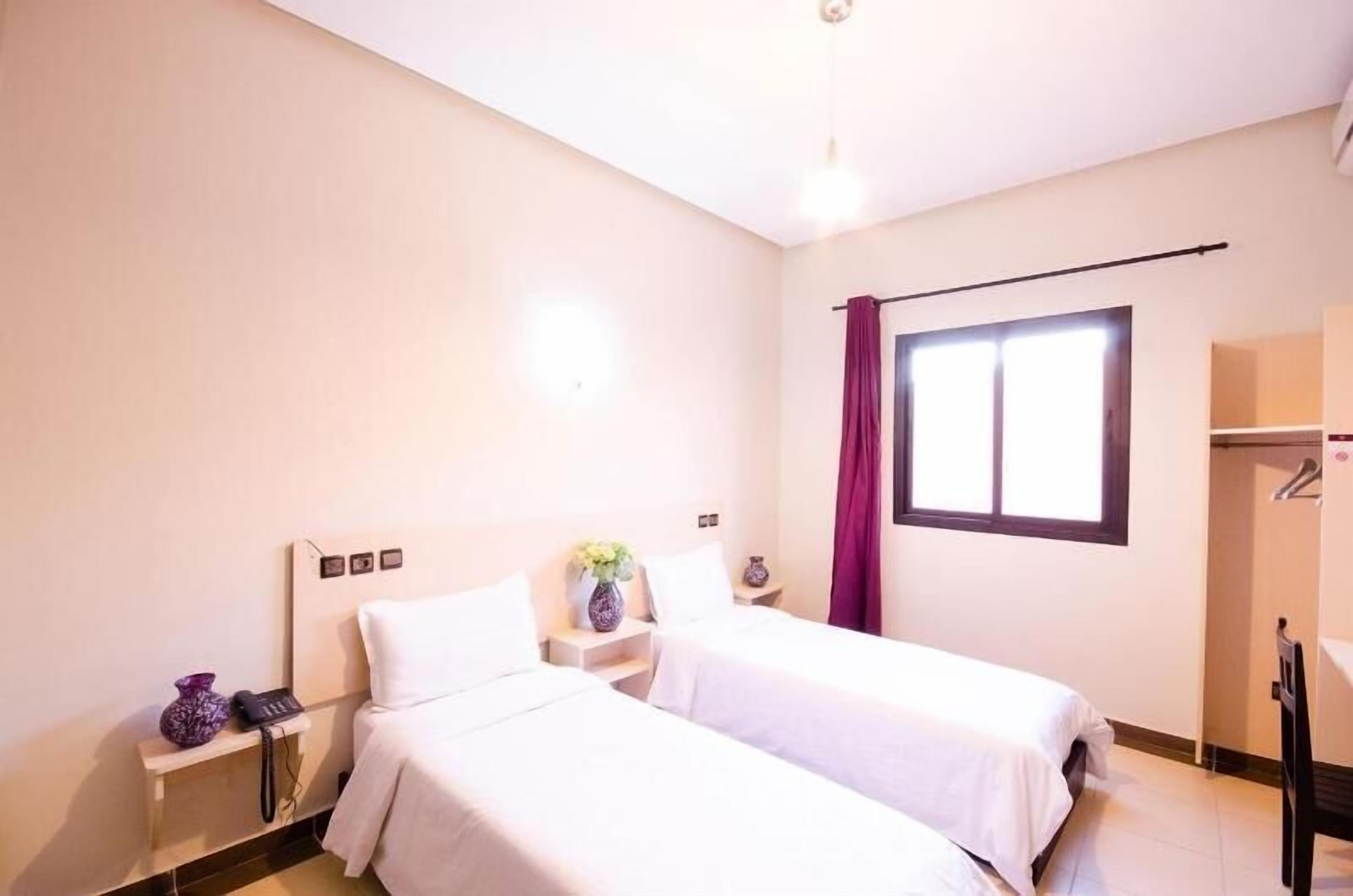 Habitación de hotel con dos camas, una mesita con un florero, un teléfono y una ventana con cortinas.