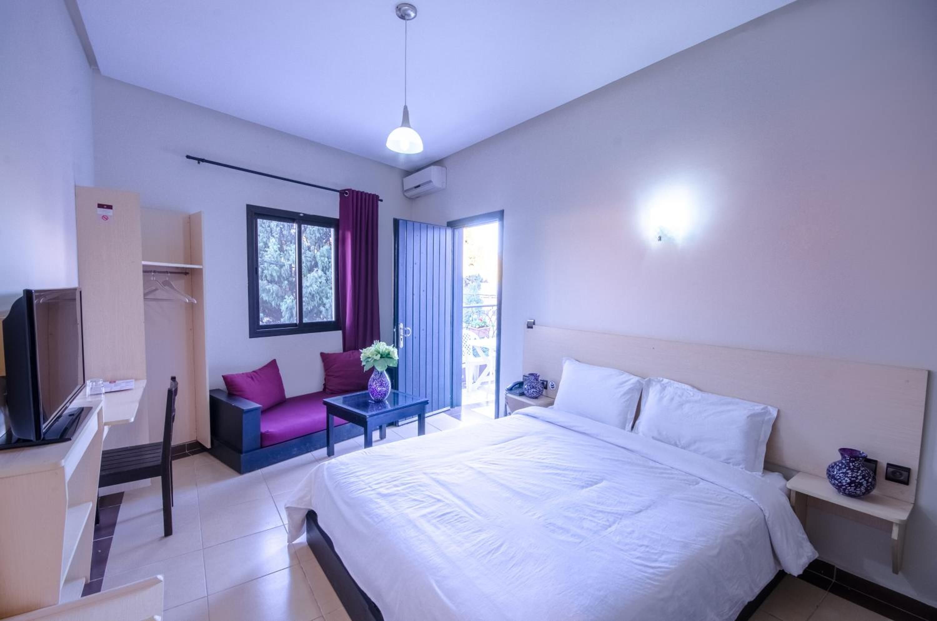 Habitación de hotel con una cama, un escritorio y una silla, un televisor, una mesita con una planta y una ventana con cortinas.