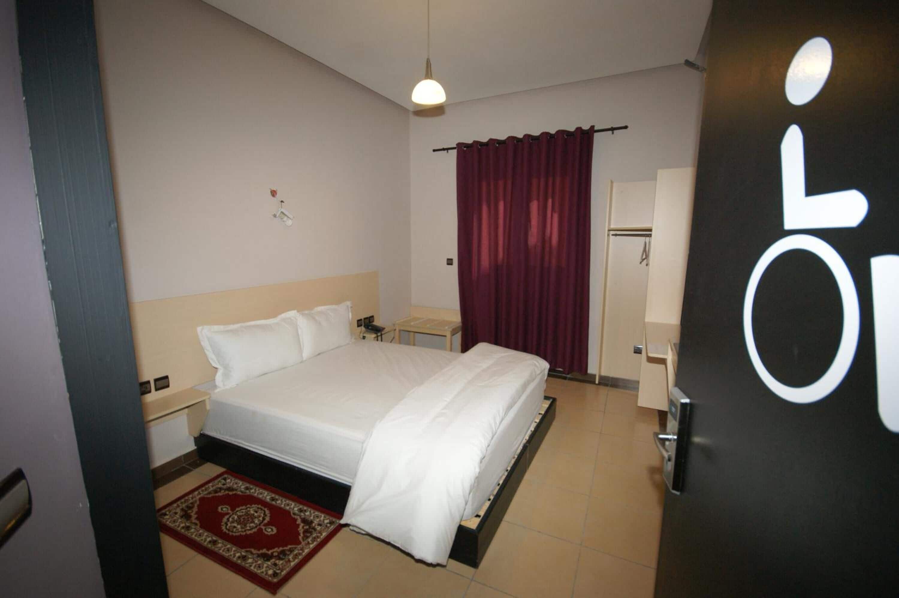 A hotel room with a bed, a nightstand, a desk, a chair, a wardrobe, and a door with a wheelchair symbol.