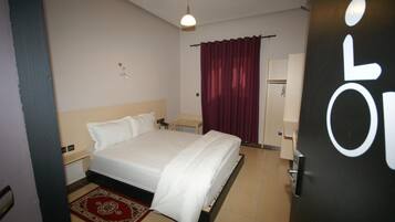 A hotel room with a bed, a nightstand, a desk, a chair, a wardrobe, and a door with a wheelchair symbol.