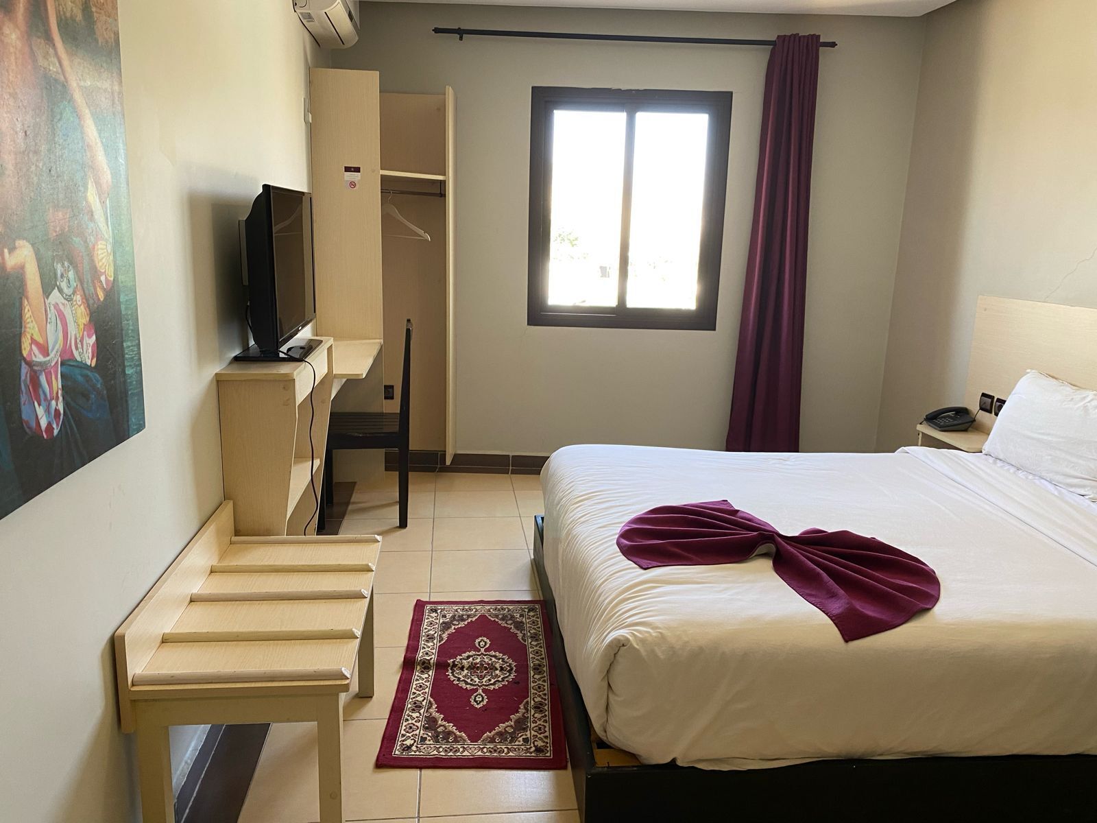 A hotel room with a bed, a desk with a chair, a television, a window with curtains, and a painting on the wall.