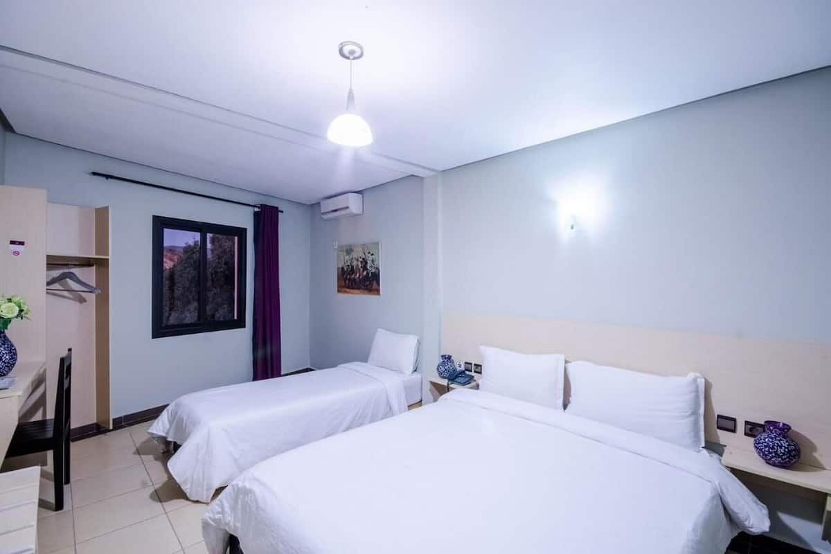 A hotel room with two single beds, a desk with a chair, a wardrobe, and a window with a view of trees.