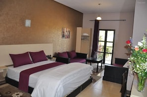 A hotel room with a large bed, a small sofa, a desk, and a chair. There is a window with curtains, a vase with flowers, and a painting on the wall.