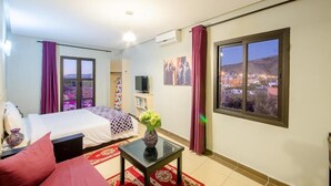 Habitación de hotel con cama, televisor, ventana que ofrece vista a la ciudad y un jarrón con flores.
