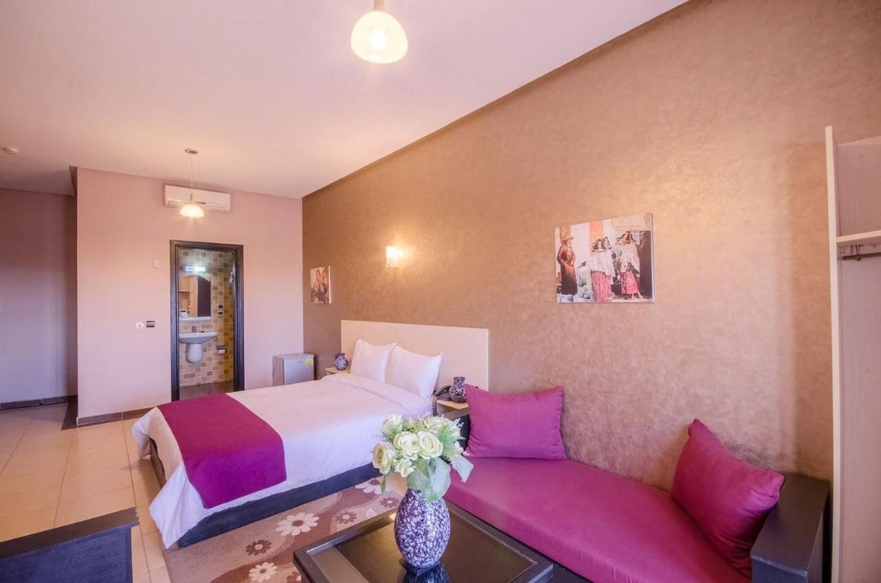 Et hotelværelse med seng, sofa, lille bord med blomstervase, natbord og vægmonteret lampe.