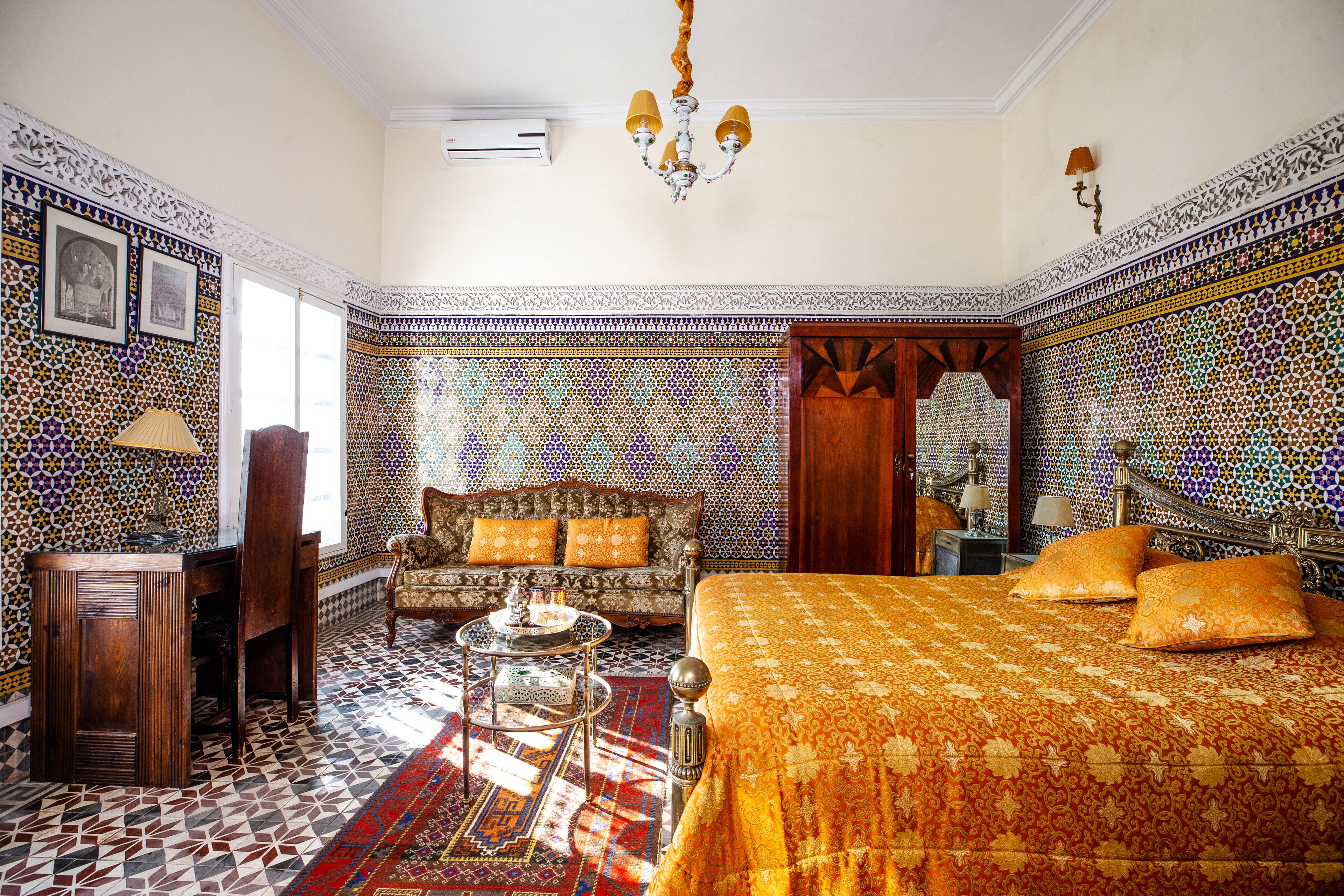 Una habitación con suelo estampado, un sofá con cojines, una cama con cubrecama estampado, un mueble de madera y una araña.