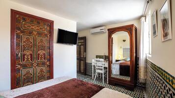 A bedroom with a large ornate door, a bed, a desk with a chair, and a television on the wall.
