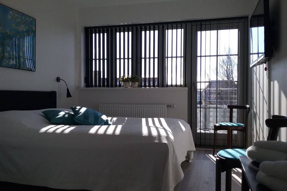 Un dormitorio con cama, radiador, ventana con persianas, una silla y un cuadro en la pared.