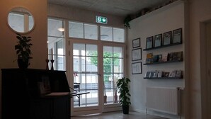 A reception area with a desk, a potted plant, a mirror, a shelf with framed pictures, and a large window with a view of the outside. - Hals Hotel (Hals)