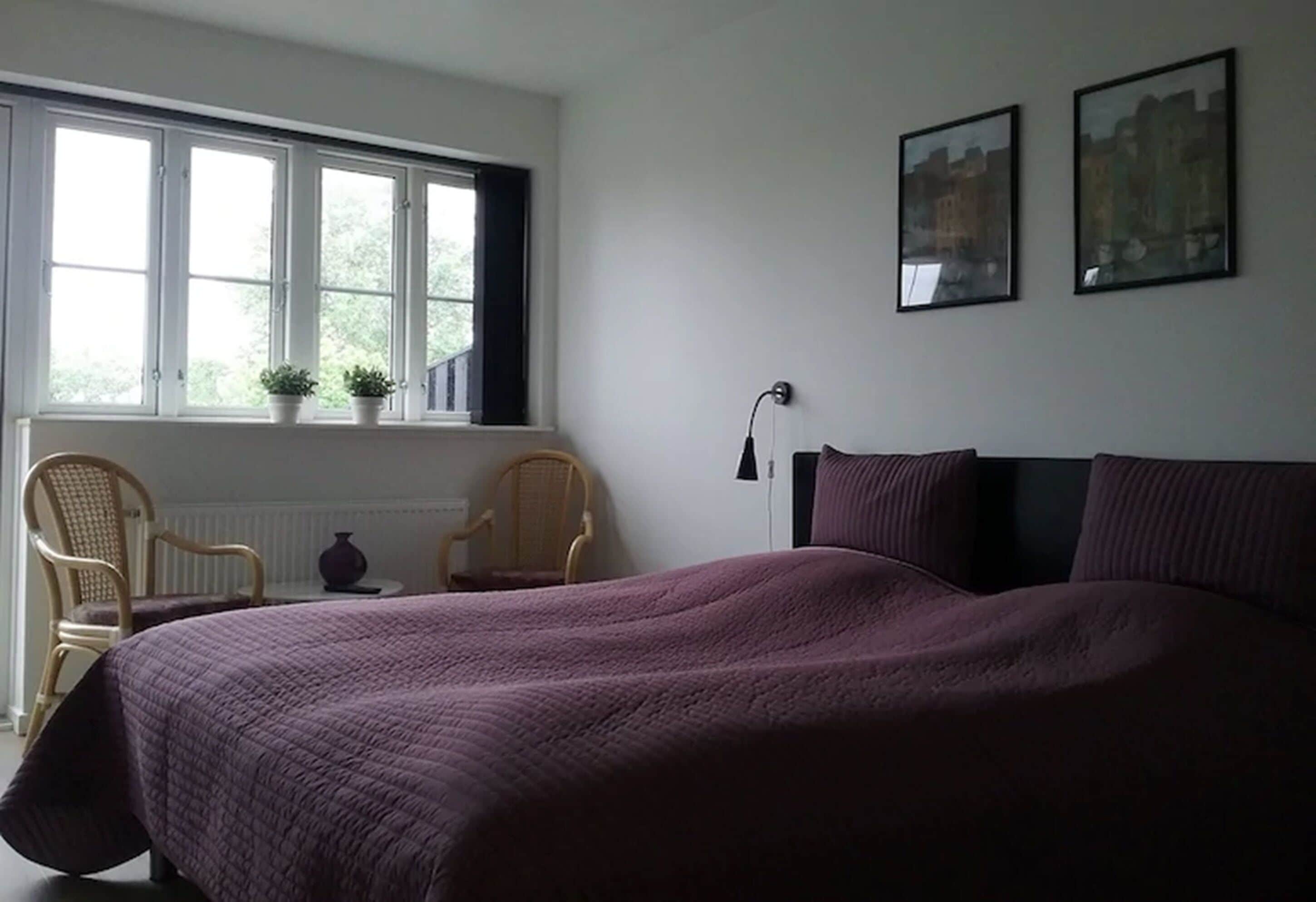 Un dormitorio con una cama grande, dos sillas, una ventana con plantas y dos cuadros en la pared.