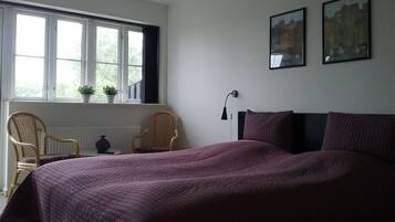 Een slaapkamer met een groot bed, twee stoelen, een raam met planten en twee afbeeldingen in lijsten aan de muur.