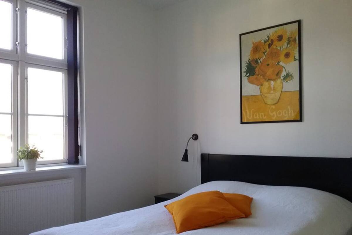 Un dormitorio con una cama, una ventana, una mesita de noche, una lámpara y un cuadro de girasoles.