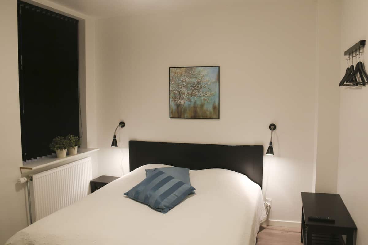 Un dormitorio con cama, mesitas de noche, un radiador y un cuadro colgado en la pared, además de una ventana con persianas.
