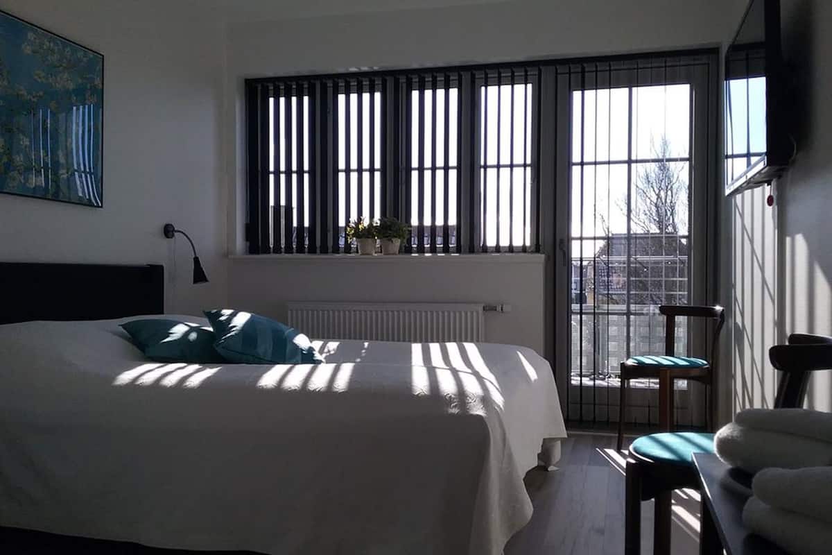 Un dormitorio con cama, ventana con persianas, radiador, una silla y un cuadro en la pared.