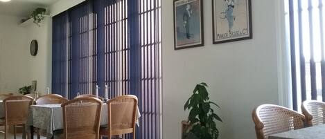 Uma área de refeições com cadeiras de vime e uma mesa, um divisório azul e dois cartazes emoldurados na parede.