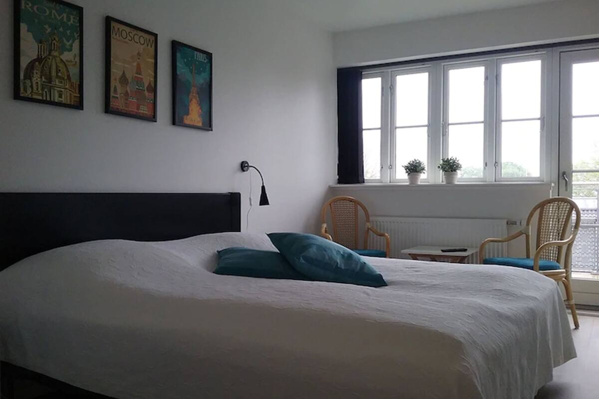 Un dormitorio con una cama grande, una ventana con cortinas, una silla, una mesita y tres carteles en la pared.