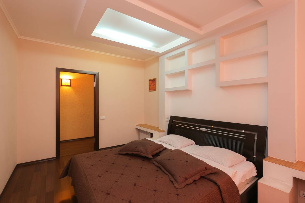 A bedroom with a bed, a bedside table, a nightstand, a ceiling light, and a door with a light fixture.