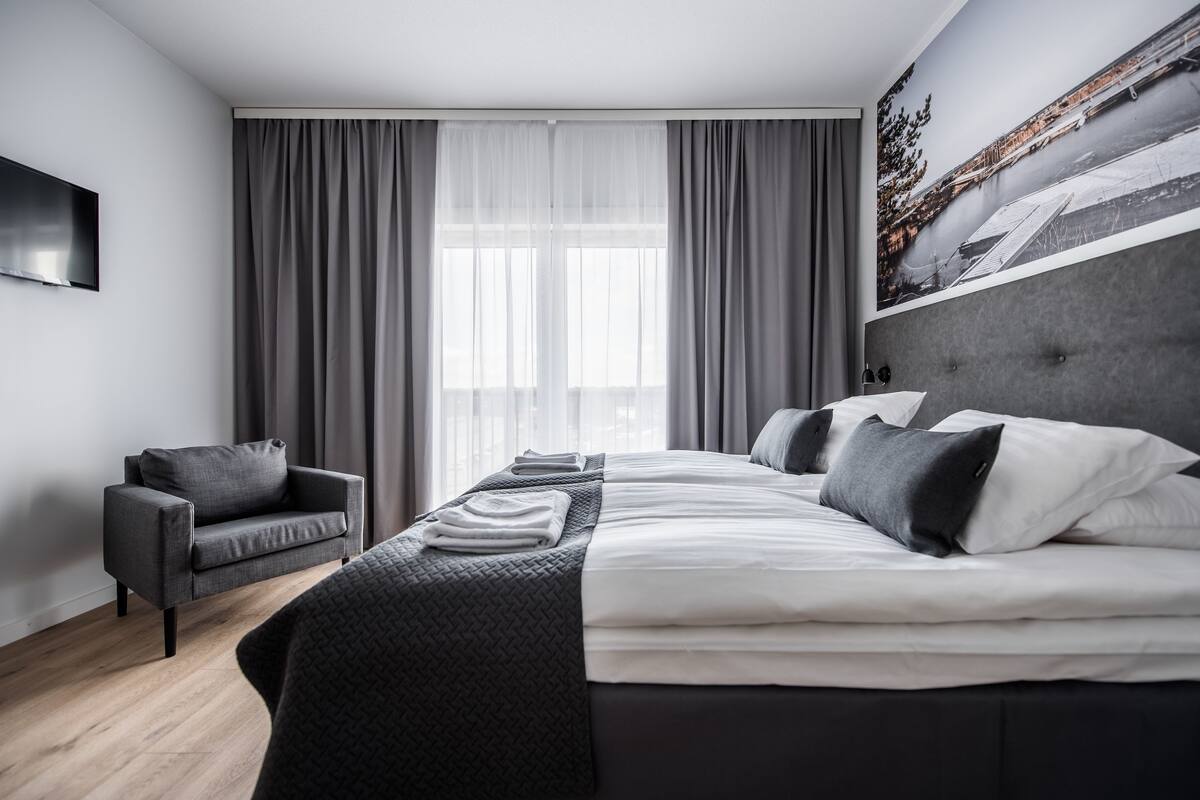 A modern hotel room with a large bed, a sofa, a flat-screen TV, and a cityscape painting on the wall.