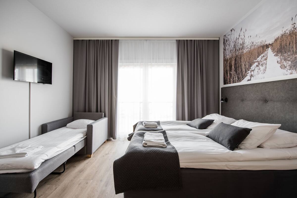 A modern hotel room with a bed, a sofa bed, a flat-screen TV, and a large window with curtains.