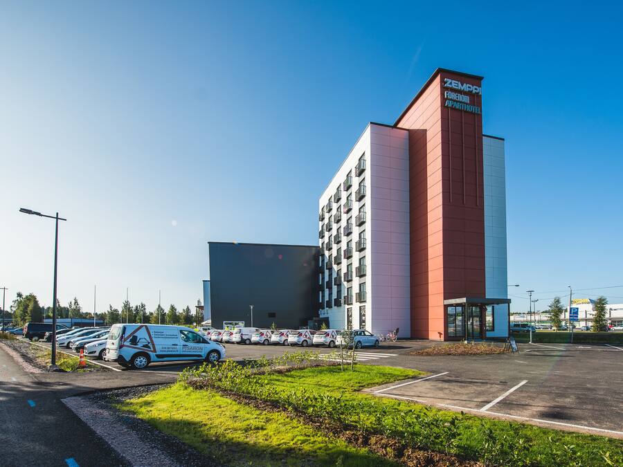 Een modern hotel met een rode en witte gevel, een parkeerplaats met verschillende auto's en een helderblauwe lucht.