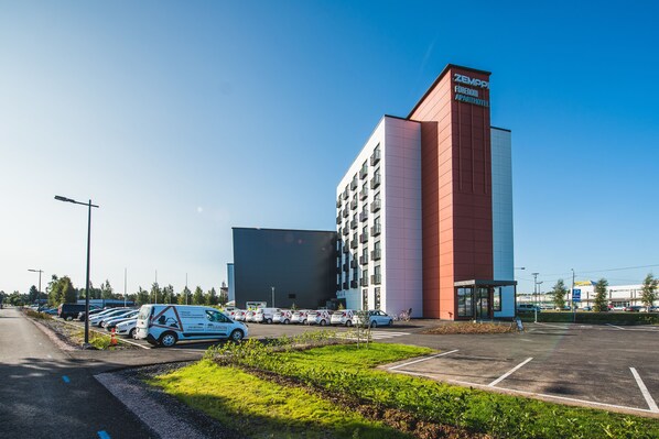 Ett modernt hotell med en röd och vit fasad, en parkeringsplats med flera bilar och en klarblå himmel.