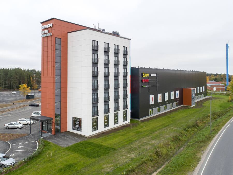 A modern hotel with a white and red facade, multiple balconies, and a parking lot.