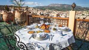 A table set for a meal with various dishes, surrounded by metal chairs and a railing, overlooking a cityscape with buildings and a mountain in the distance. - Riad Fes Maya (Fes)