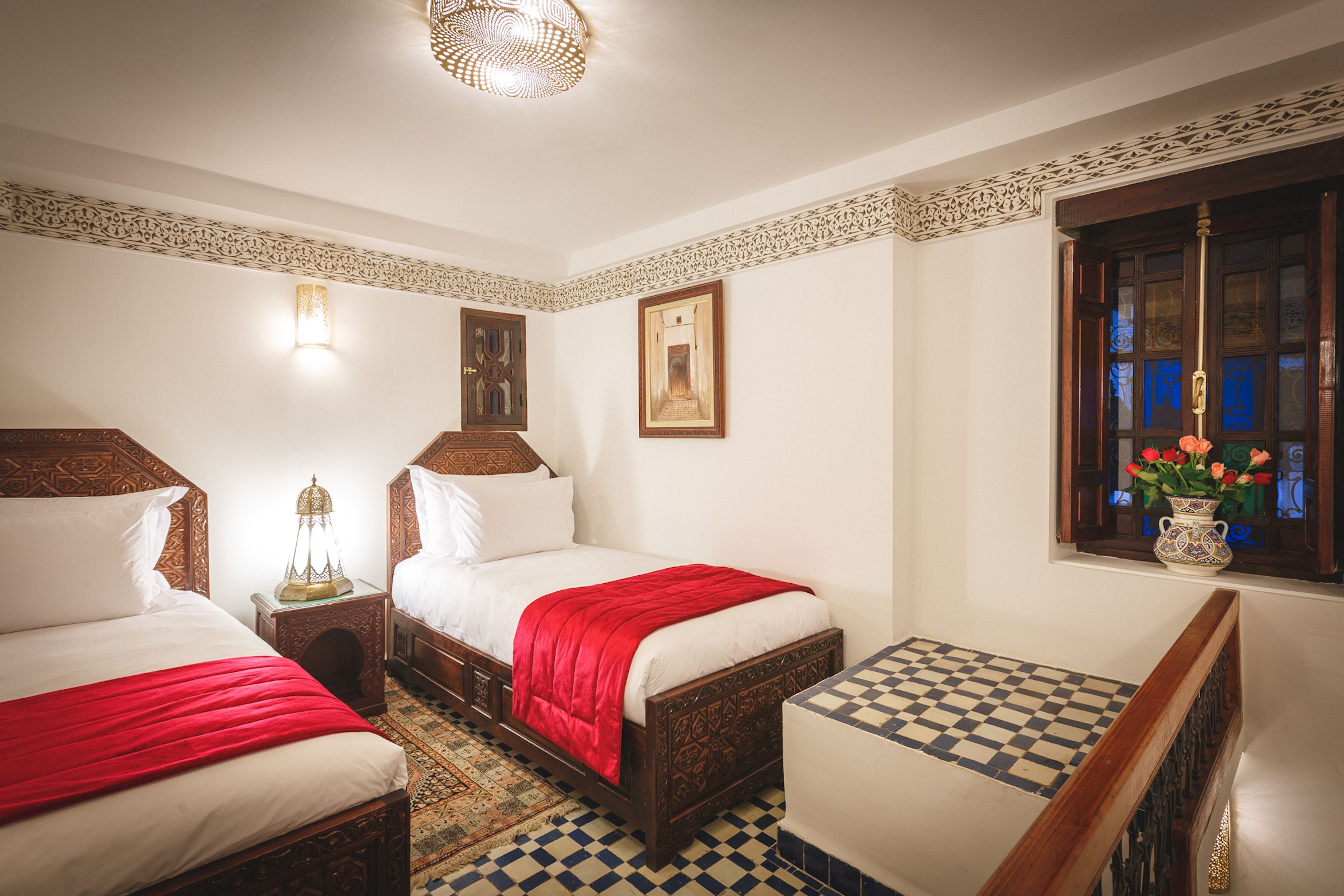 Ein Hotelzimmer mit zwei Betten, einem Schachbrettmuster-Boden, einem Zierfenster und einem Nachttisch mit Lampe.