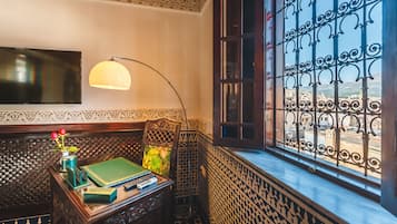 Um quarto com uma escrivaninha de madeira, livros, um vaso com flores, uma luminária e vista através de uma janela com grades.